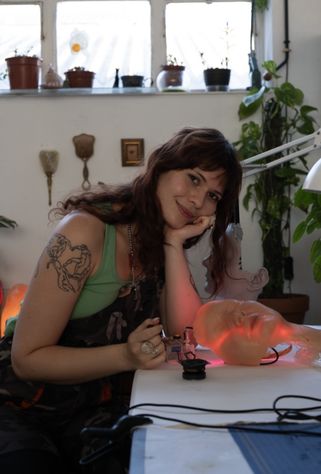 Manuela in her studio, chin resting on her hand, beside a silicone head glowing red on the worktable