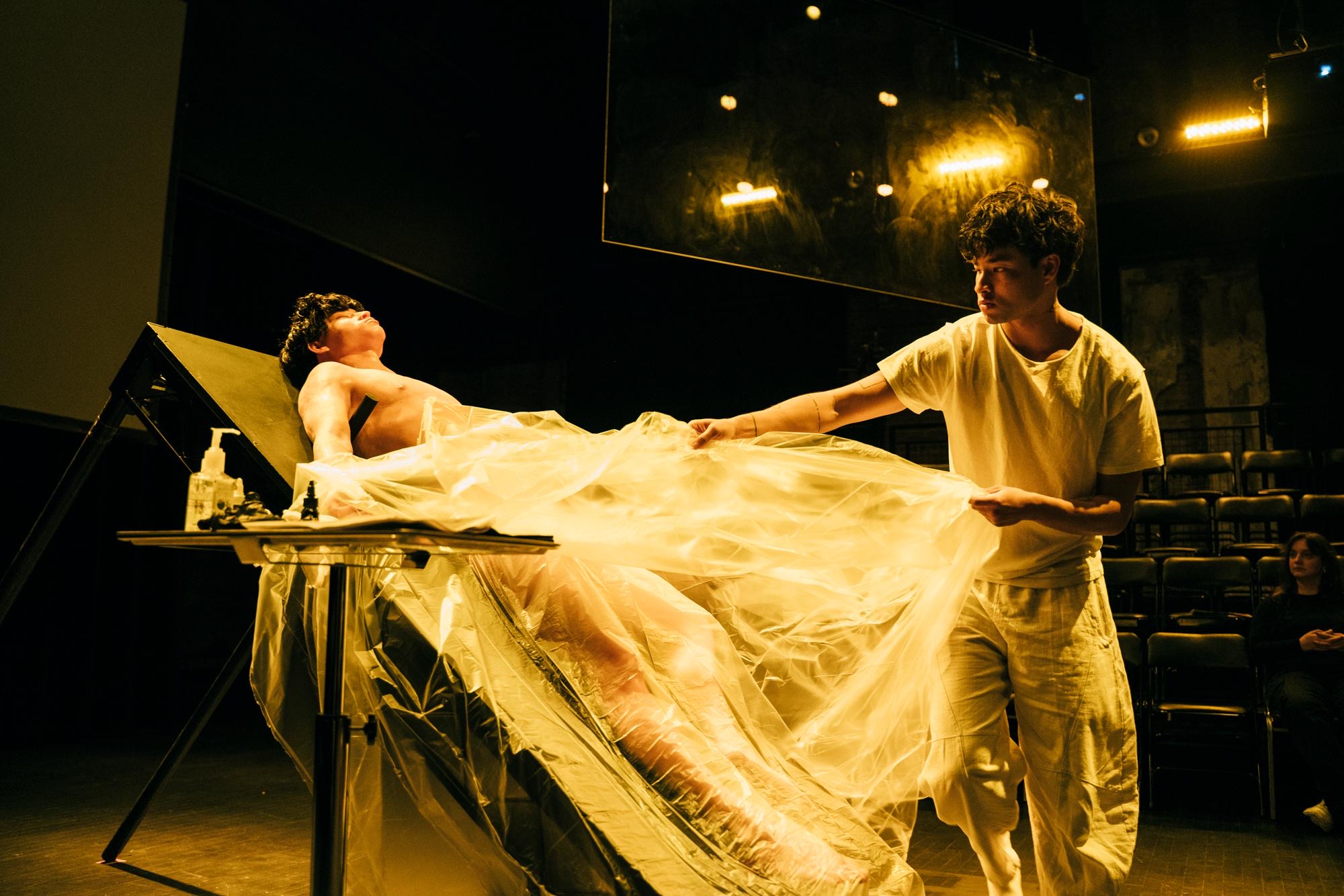 A performer lies back on a tilted table under clear plastic sheeting while another pulls the plastic away