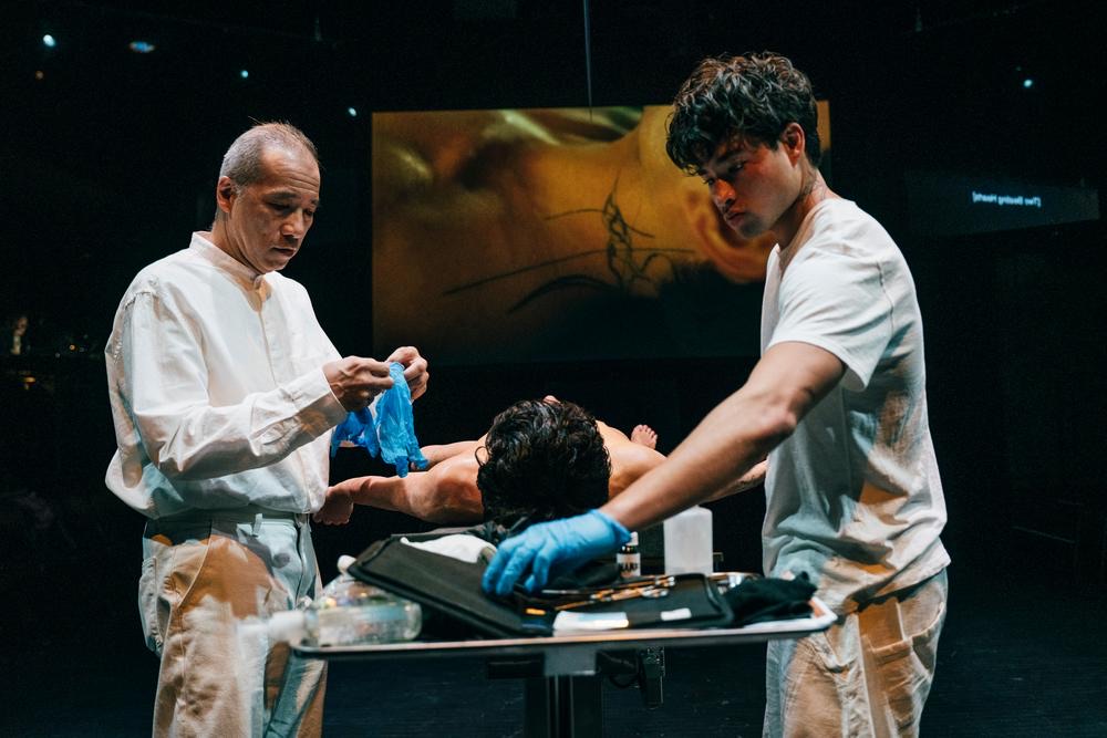 Two performers pull on blue gloves over a body lying on the table, a projection behind them