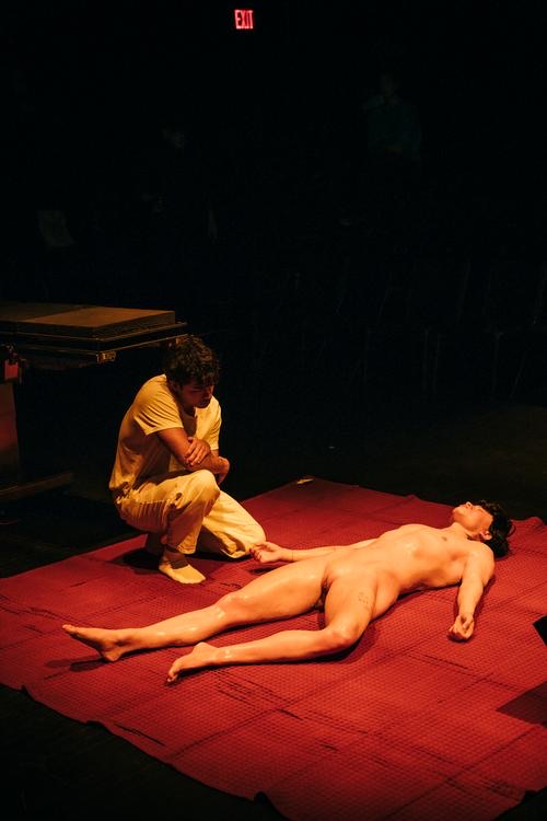 A performer crouches beside a full-body silicone replica lying on a red rug