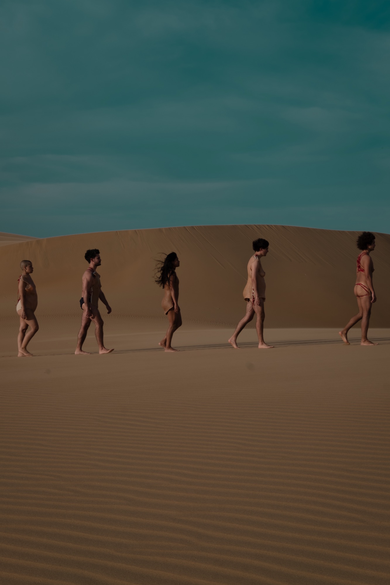 Five performers in silicone body skins walk in a line across a sand dune under a dark teal sky