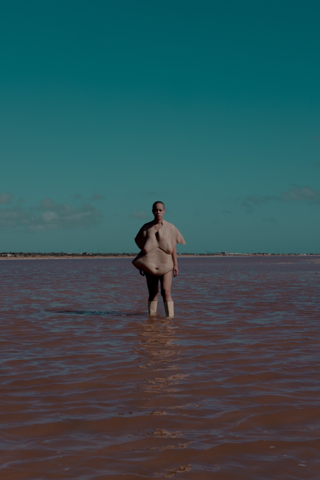 A performer wearing a silicone torso skin and white boots stands alone in shallow pink water