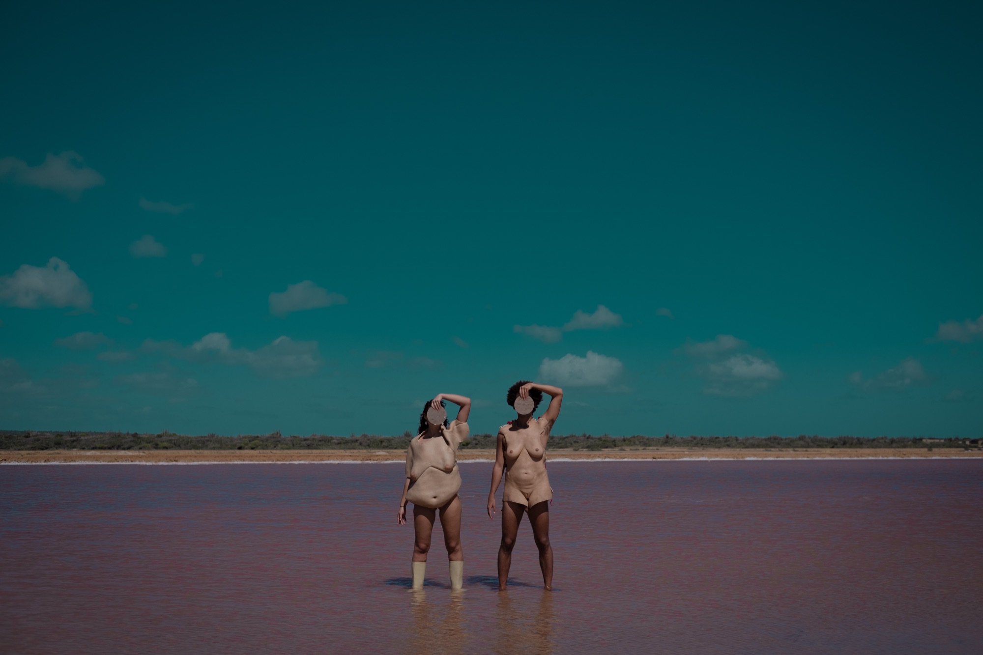 Two performers in silicone body skins stand in a pink salt lake, each holding a round mirror over their face
