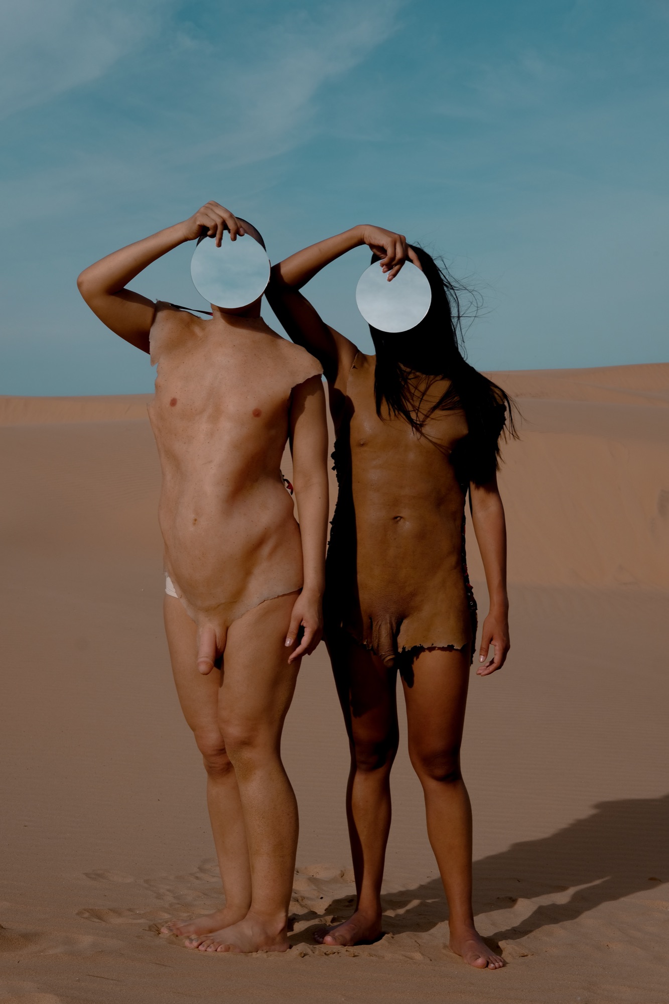 Two performers in silicone body skins stand on a dune, round mirrors hiding their faces