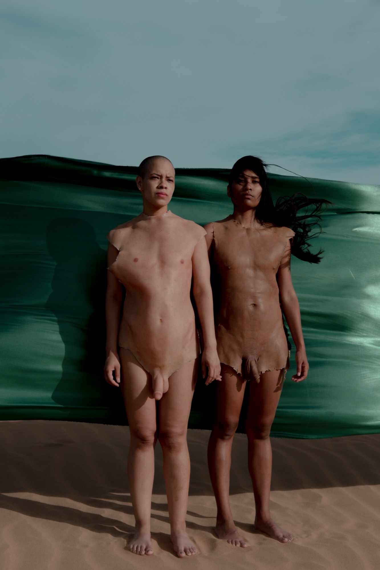 Two performers in silicone body skins stand on the sand in front of a billowing green fabric